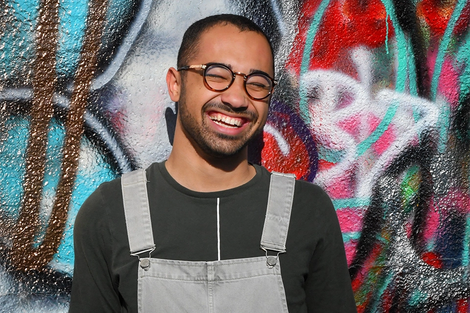 Man, an LGBTQ+ influencer, smiling in overalls in front of graffitid wall