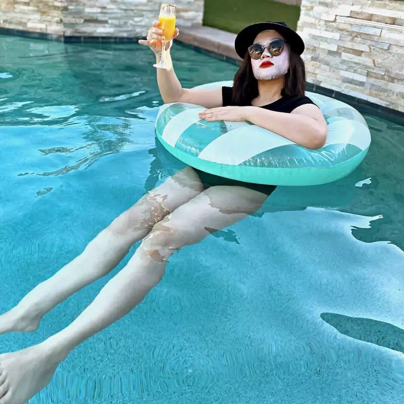 Woman floating in a pool ring enjoying a mimosa with a cosmetic facial mask on