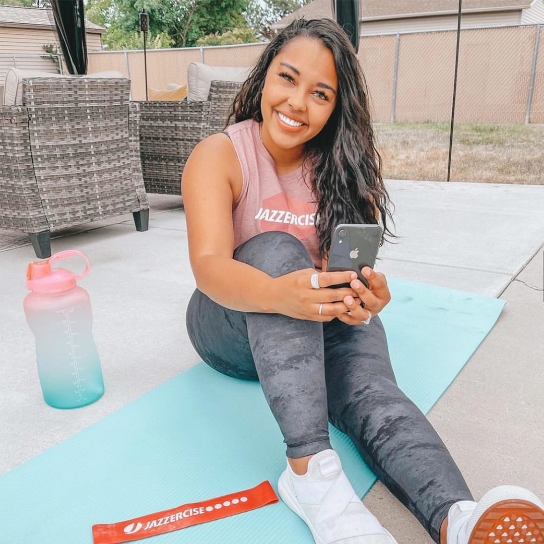 woman smiling and holding a phone while sitting on a yoga mat outside