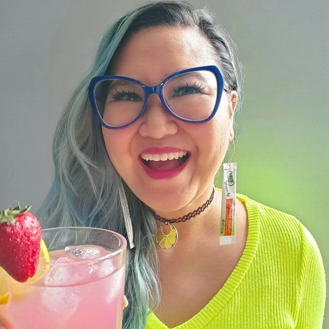 A woman with glasses and True Citrus earrings holding a glass of pink True Citrus drink