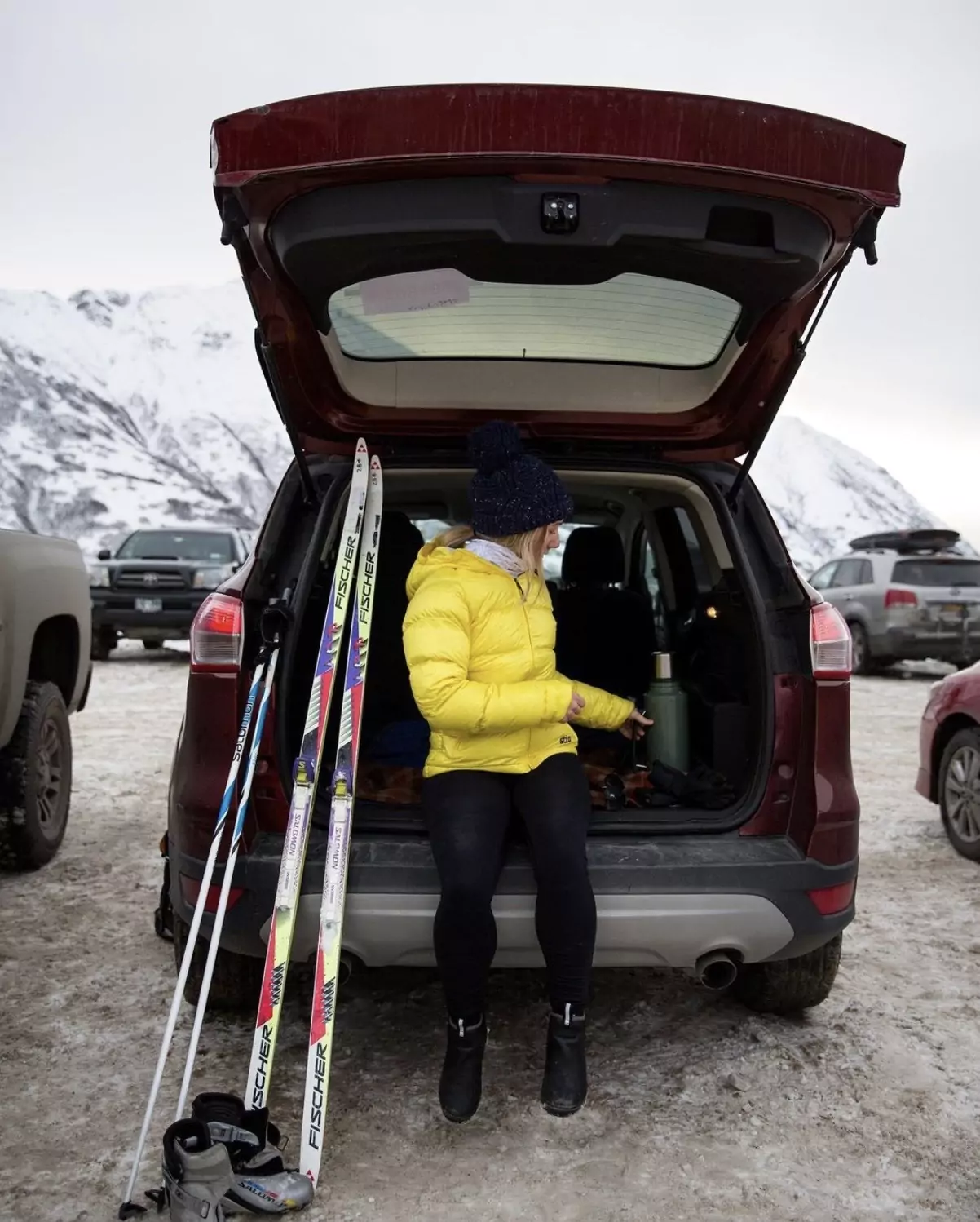 A woman in a yellow puffy jacket sitting in an open SUV trunk net to skiis